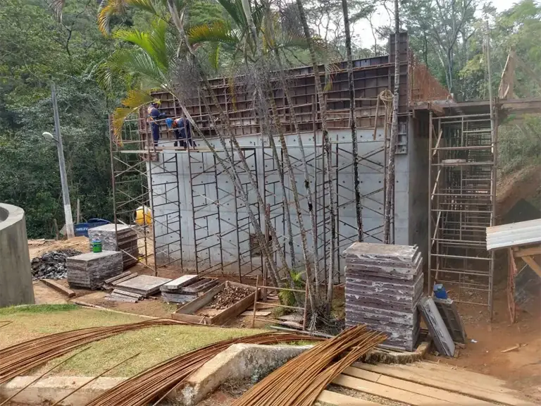 Parede alta com forma metálica para ponte e andaimes em obra cercada de vegetação